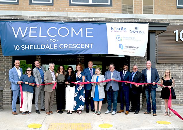 A group of smiling people cutting a large ribbon outside a new apartment building.